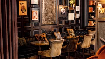 Eine schummrig beleuchtete Bar mit Ledersitzmöbeln, gerahmten Kunstwerken an den Wänden und einem Bücherregal im Hintergrund.