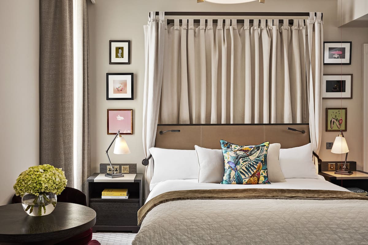 A hotel room with a large bed, a nightstand with a lamp, a round table with a vase of flowers, and a view of the city through the window.