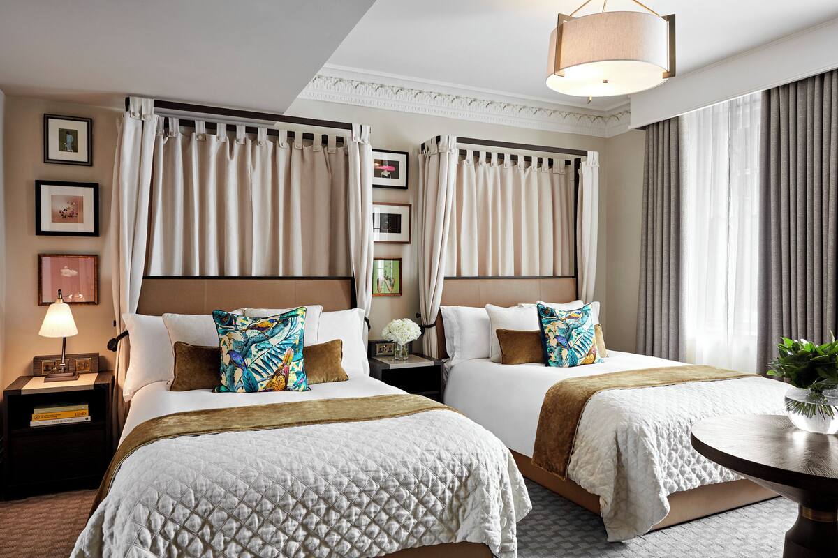 A hotel room with two beds, a bedside table with a lamp, a small table with a plant, and a large window with curtains.