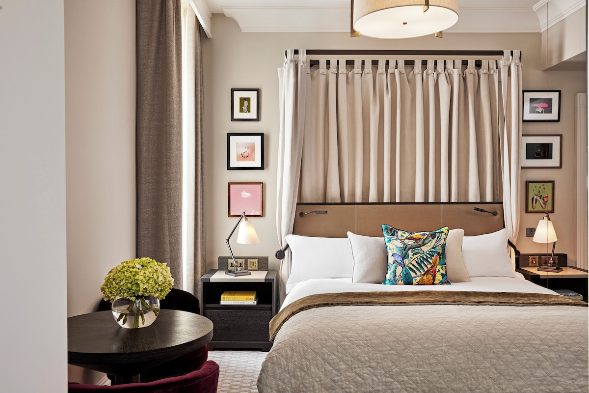 A hotel room with a large bed, a bedside table with a lamp, a small round table with a vase, and a view of the city through the window.