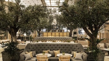 A luxurious indoor seating area with a large tufted sofa, round tables, and wicker chairs, surrounded by potted plants and a high ceiling with a skylight.