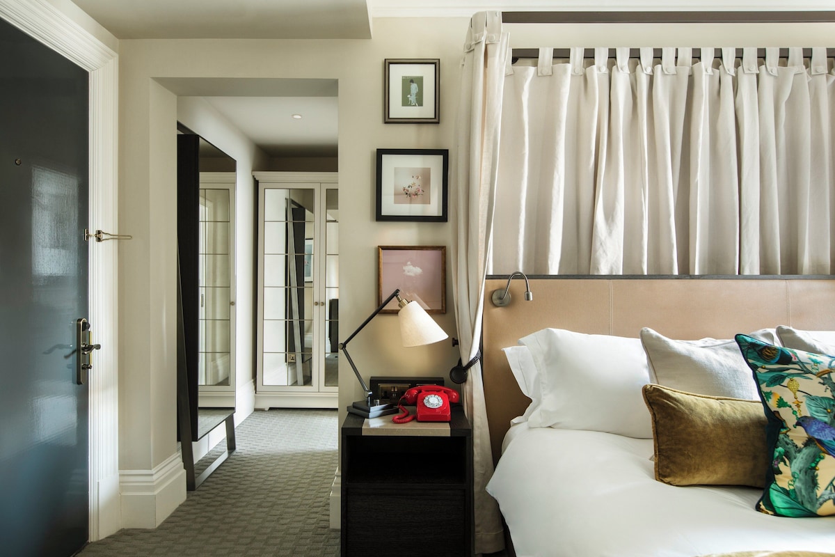 A hotel room with a bed, a nightstand with a lamp and a red telephone, a window with curtains, and framed pictures on the wall.