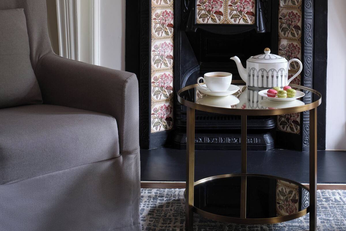 A room with a fireplace, a white mantelpiece, a sofa, a table with a teapot and macarons, and a television mounted above the fireplace.