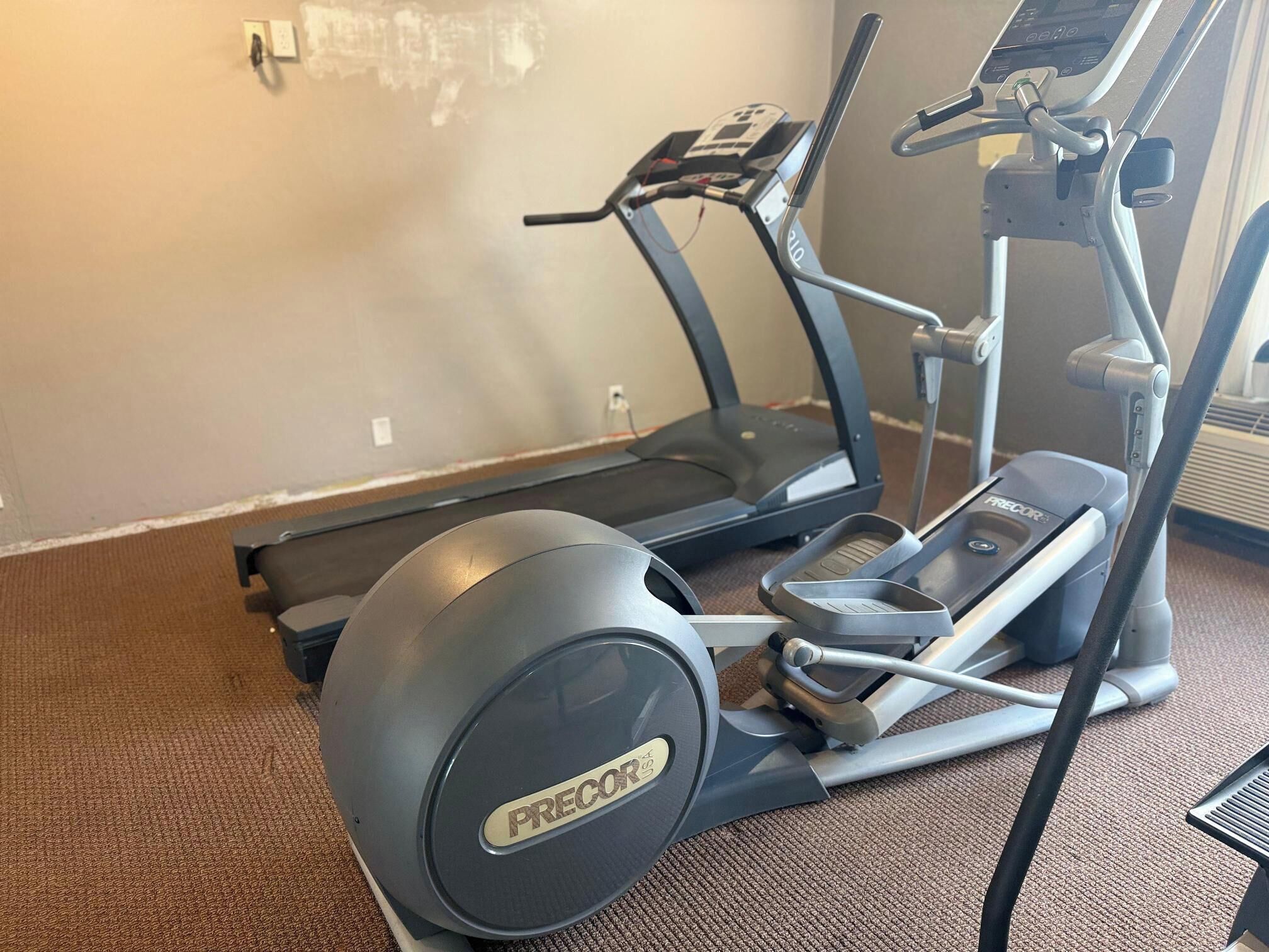 A stationary bike and a treadmill in a room with carpet flooring and a plain wall.
