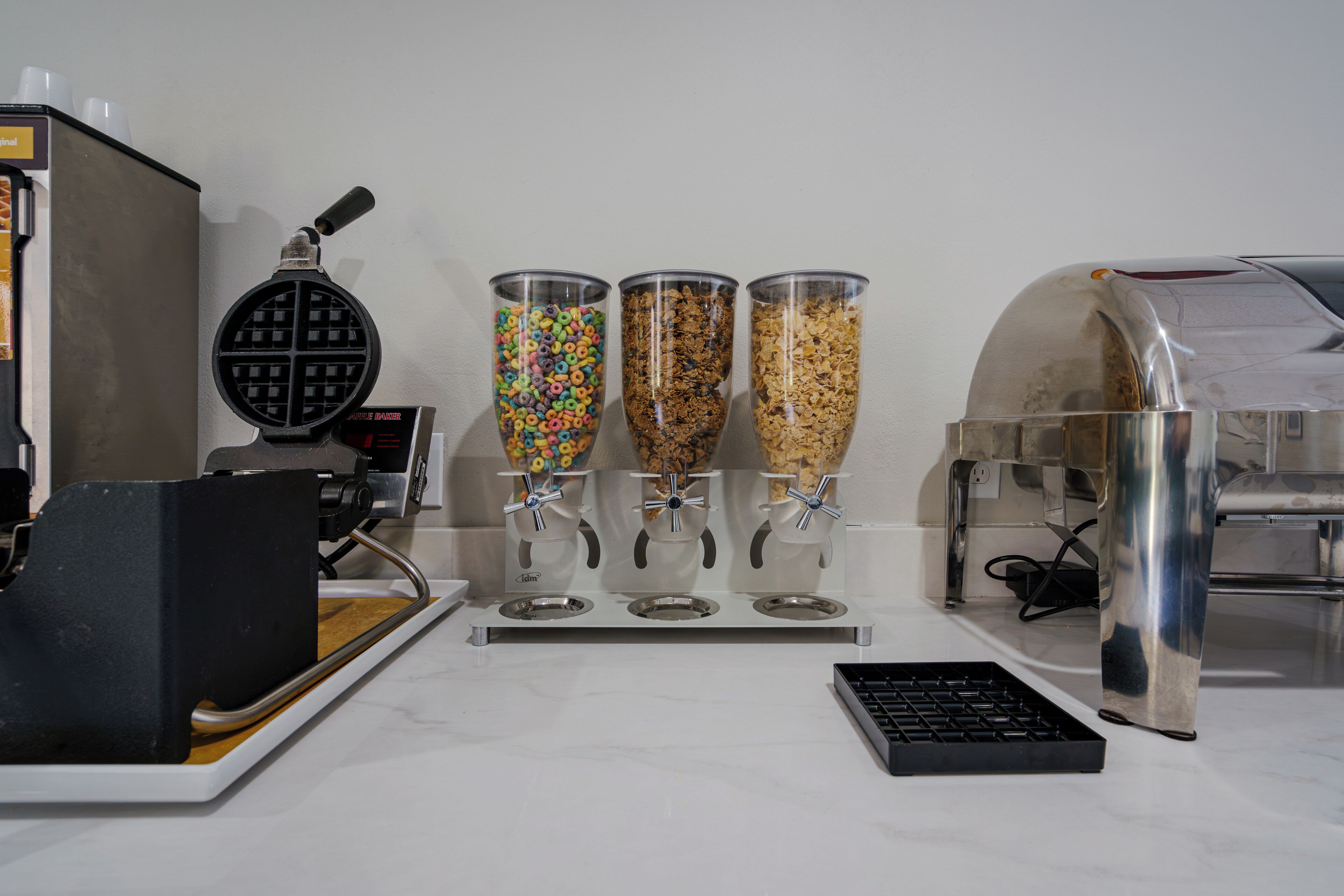 A waffle maker, three dispensers with different cereals, and a coffee machine on a kitchen counter.