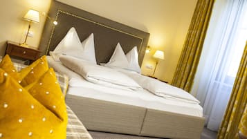 A neatly made bed with white linens and pillows, a headboard with a decorative trim, two bedside lamps, and a window with yellow curtains.