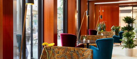 Un moderno vestíbulo de hotel con paneles de madera, un ventanal grande y una zona de estar con tapicería estampada y un jarrón con flores amarillas.