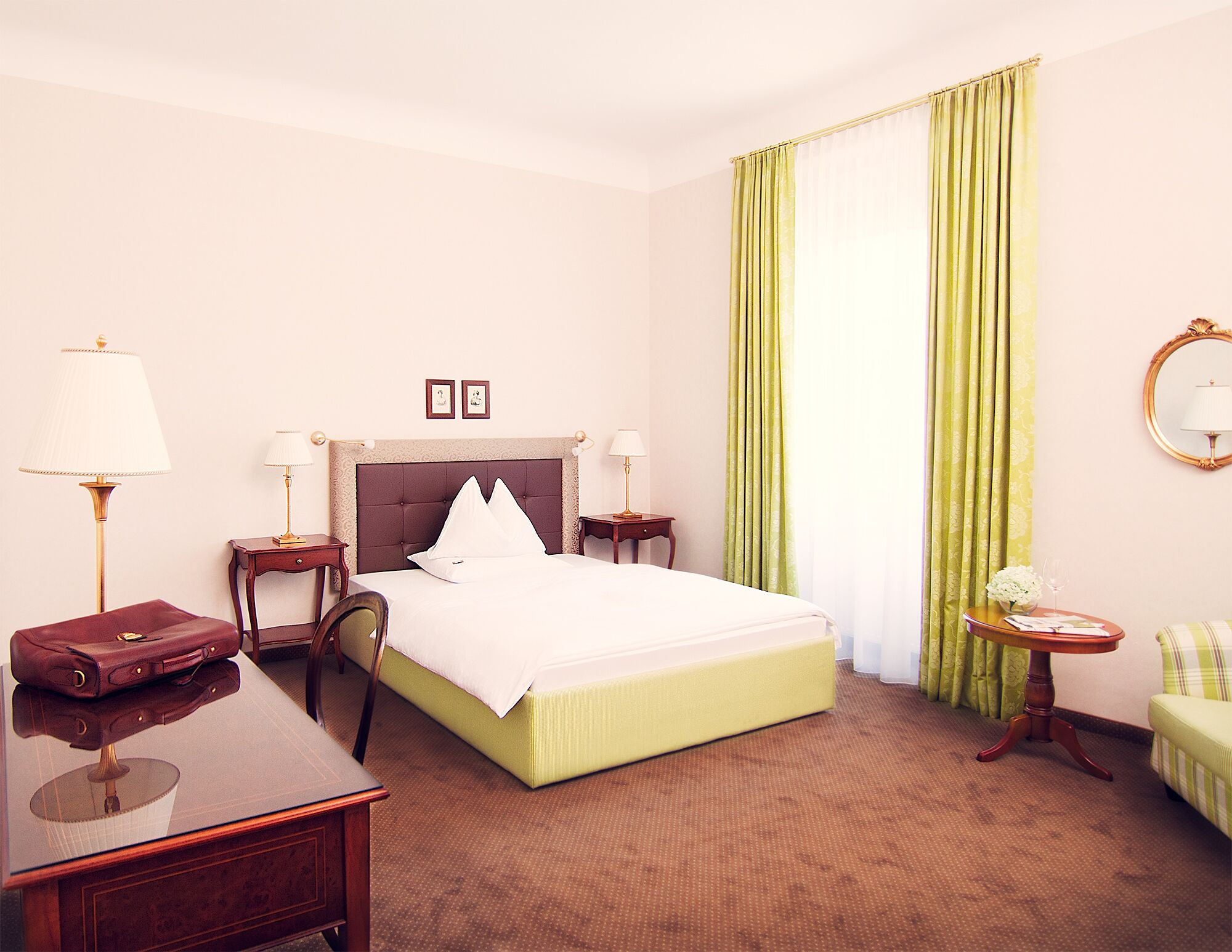 A hotel room with a large bed, two bedside tables with lamps, a desk with a chair, a small round table with a vase and glass, and a green armchair.