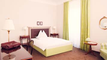A hotel room with a large bed, two bedside tables with lamps, a desk with a chair, a small round table with a vase and glass, and a green armchair.