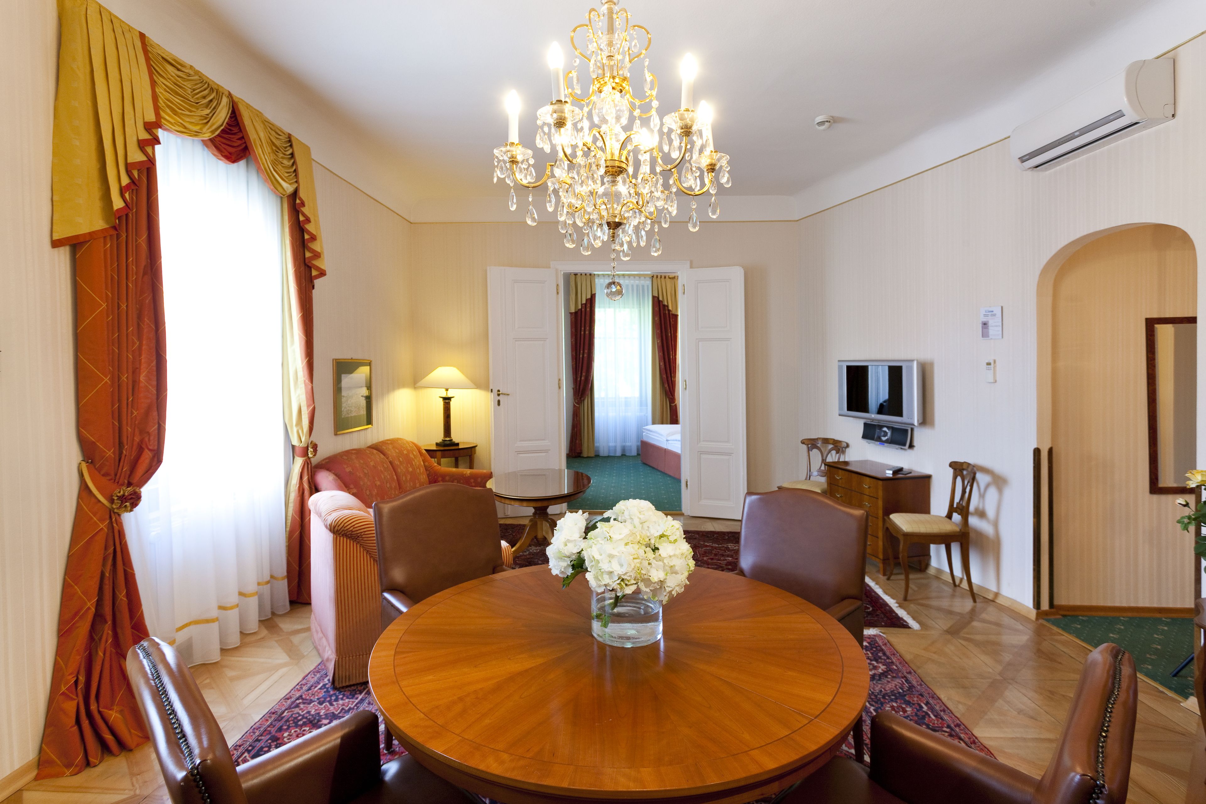 A hotel room with a round dining table, chairs, a chandelier, a television, and a sofa.