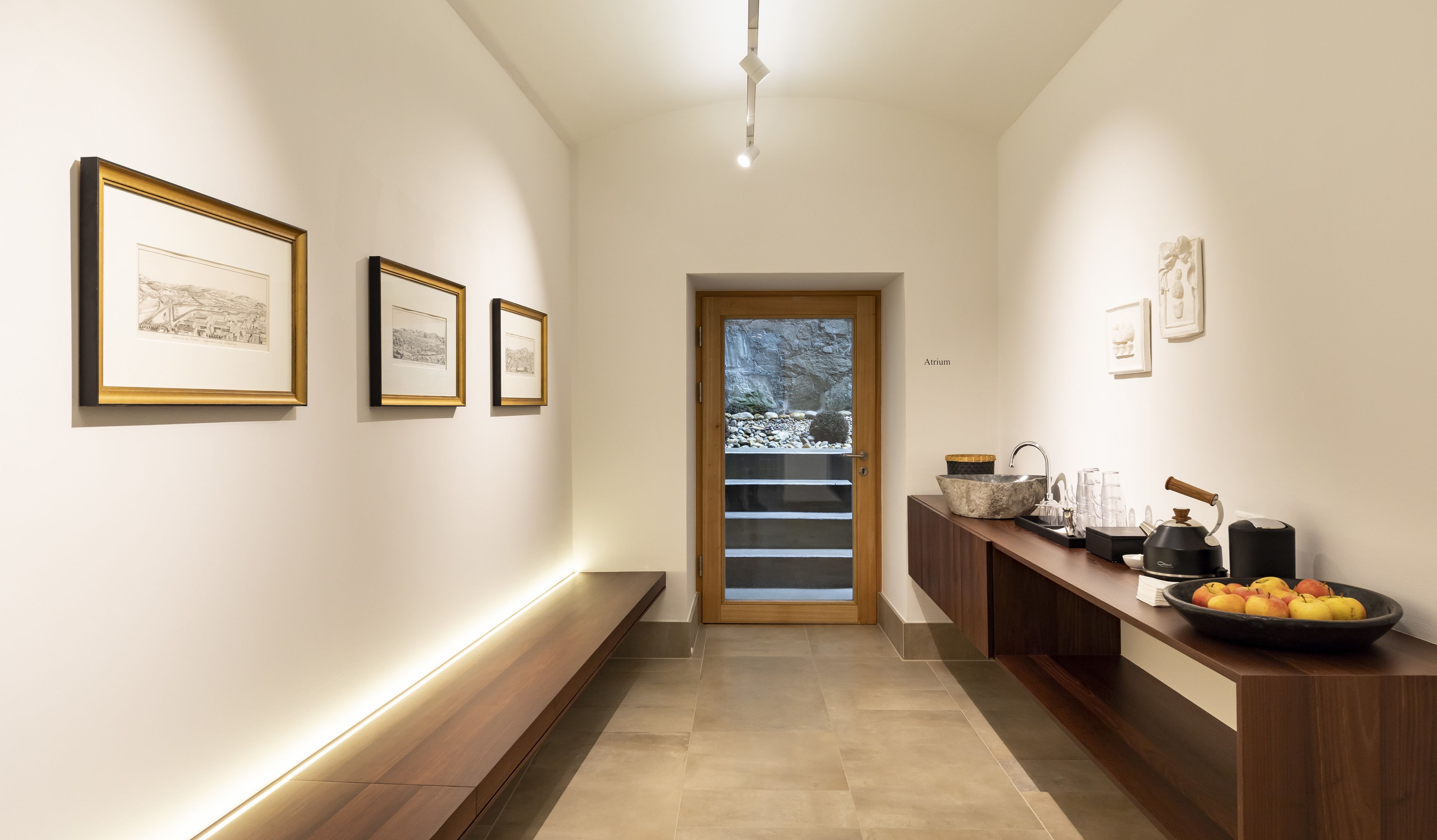 A hallway with framed artwork on the walls, a wooden door with a glass panel, and a kitchen area with a sink and countertop.