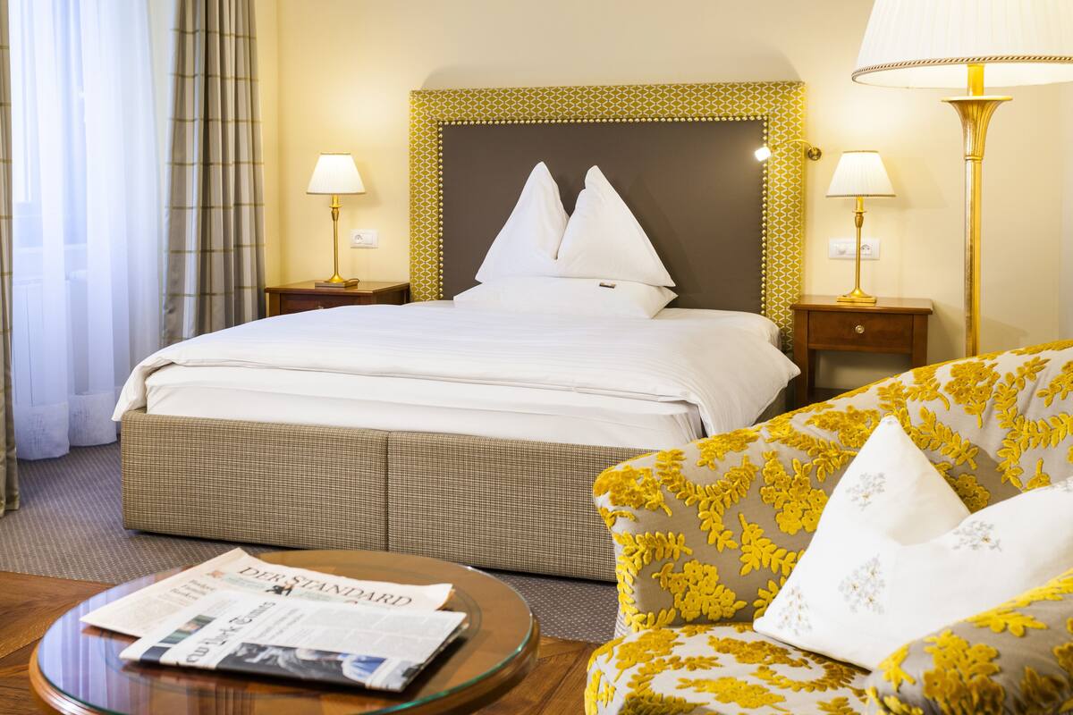 Ein Hotelzimmer mit einem großen Bett, zwei Nachttischen mit Lampen, einem Sofa mit gelbem Blumenmuster und einem runden Glastisch mit einer Zeitung.