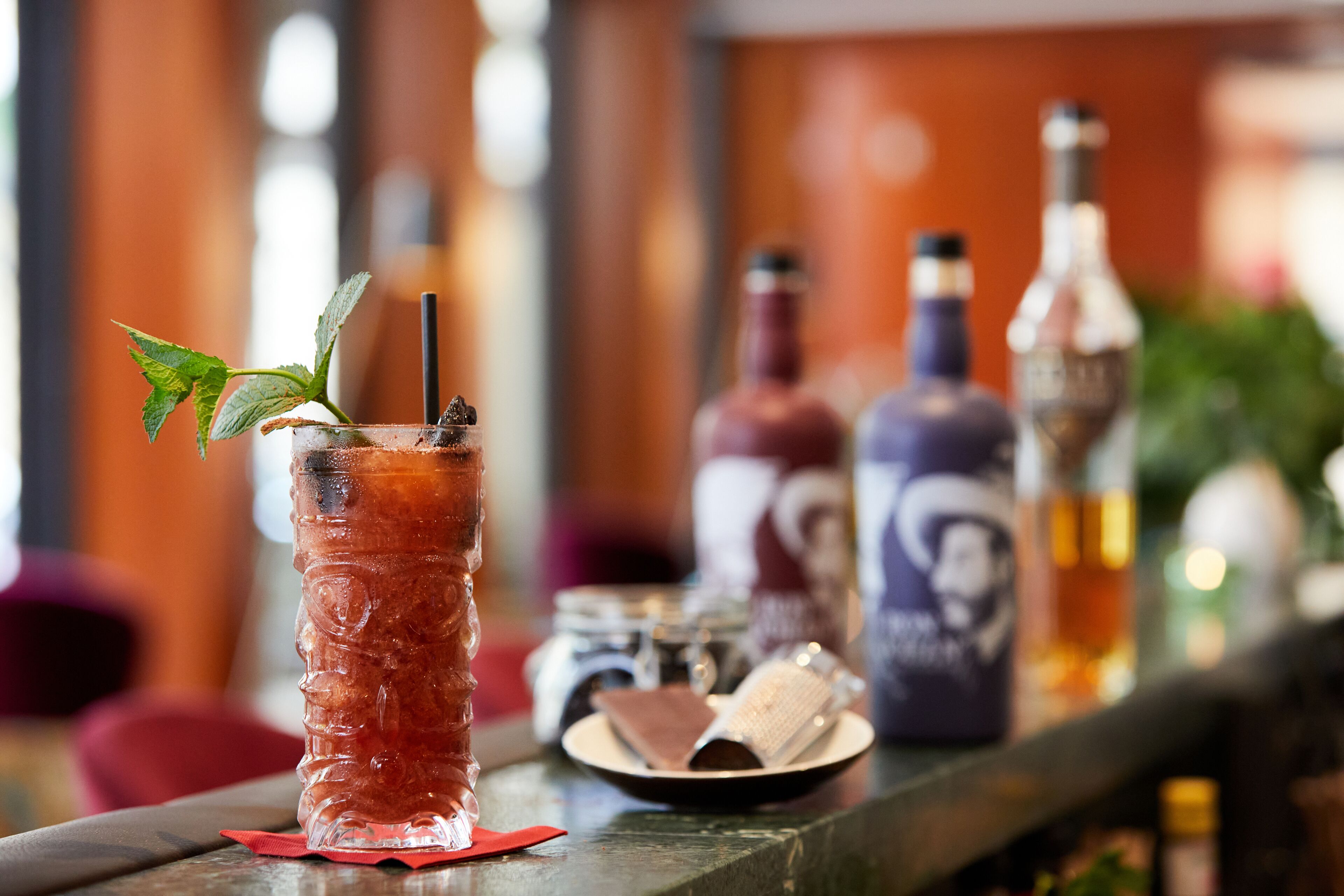 A bar setting with a tall glass of a red beverage garnished with a mint leaf and a straw, accompanied by various bottles and a small plate with a dessert.
