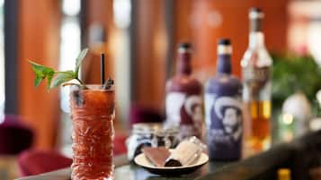 Un bar con un vaso alto de una bebida roja adornado con una hoja de menta y una pajita, acompañado de varias botellas y un plato pequeño con un postre.