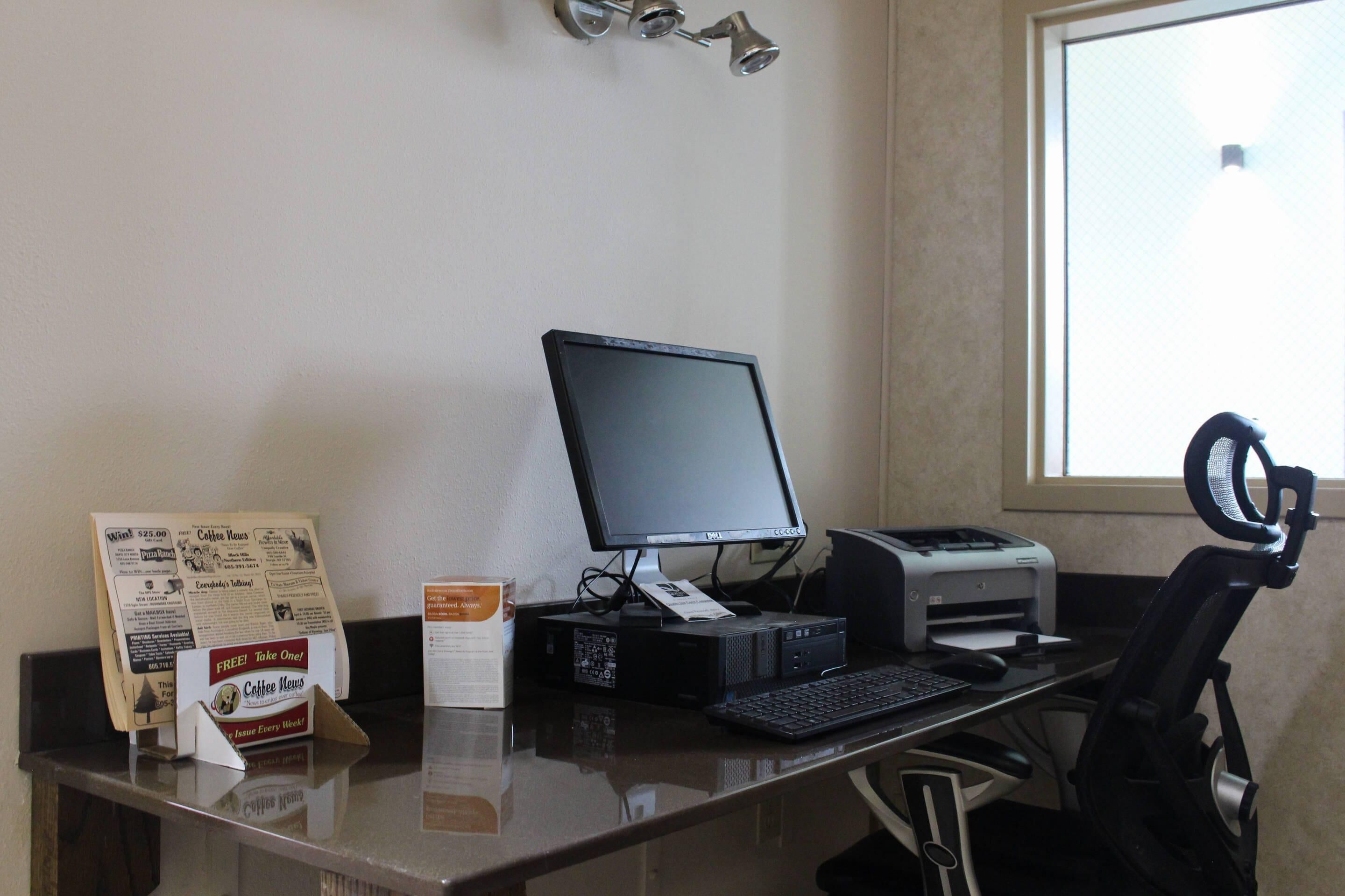 A computer desk with a monitor, keyboard, and printer. There is a chair and a window with blinds.
