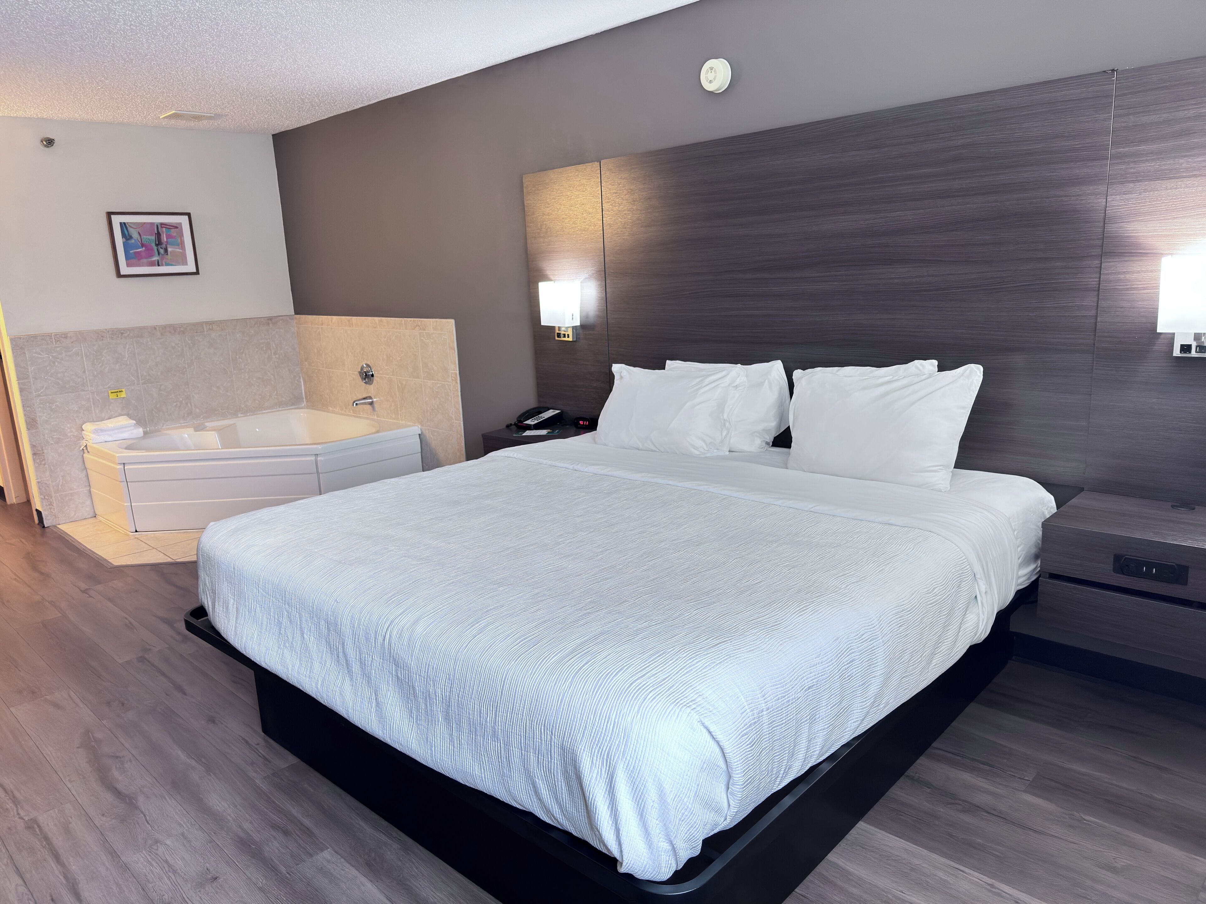 Quarto de hotel moderno com uma cama grande, mesa de cabeceira, luminária na parede e banheiro com banheira e pia.