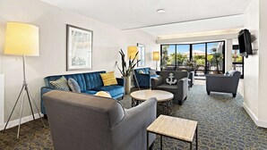 Habitación de hotel moderna con un sofá azul, un sillón gris, una mesa de centro redonda y un ventanal con vistas al exterior.