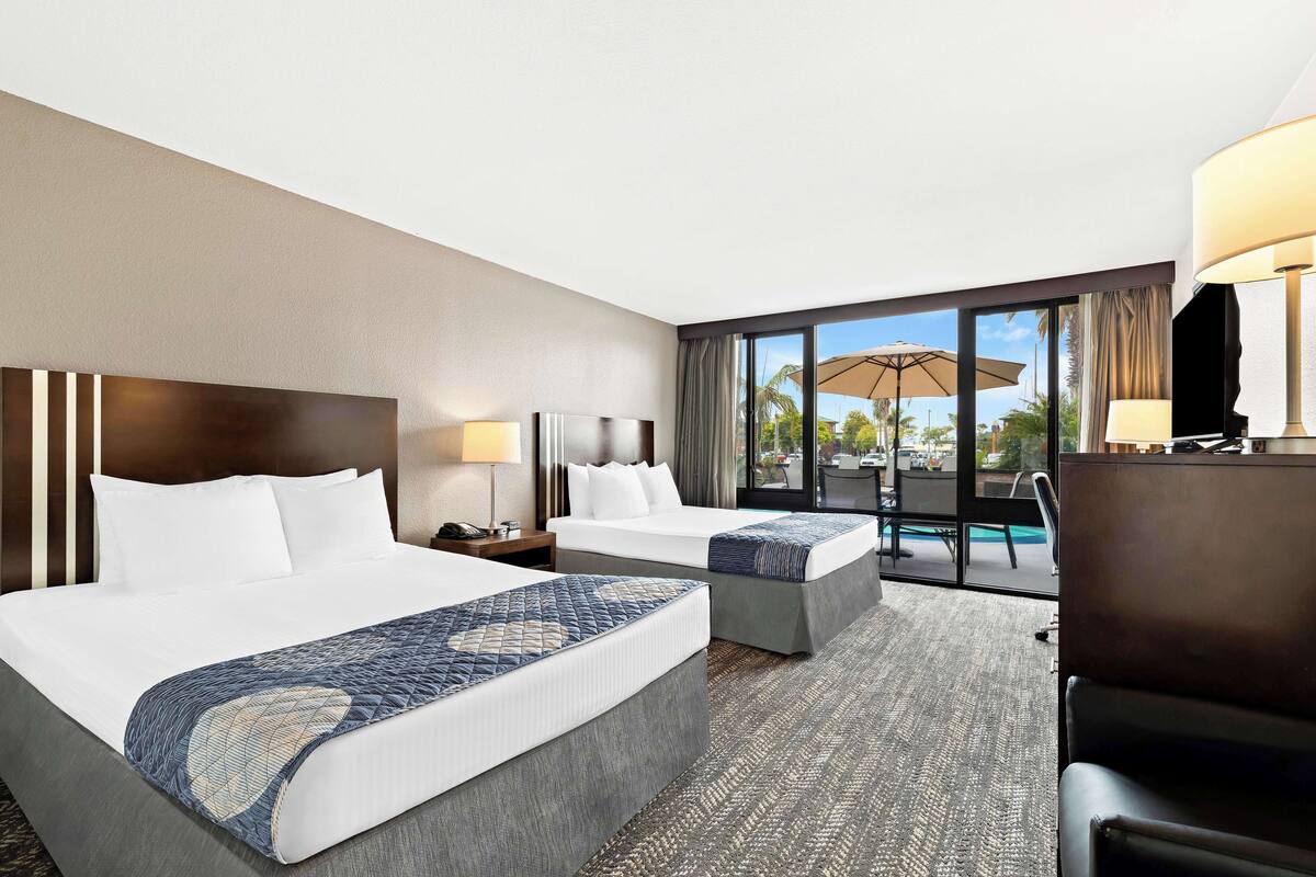 A hotel room with two beds, a desk, a chair, and a large window with a view of the outdoors.