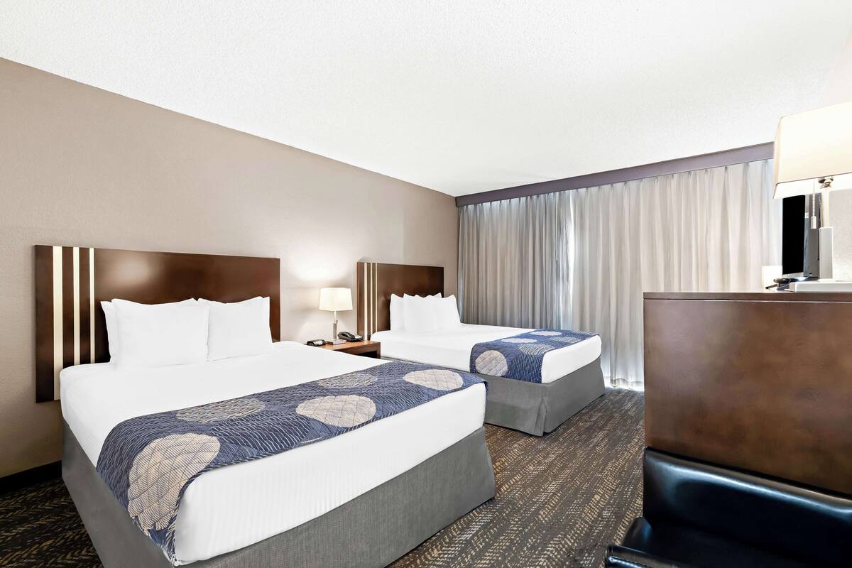 A hotel room with two beds, a desk, a lamp, and a large window with curtains.