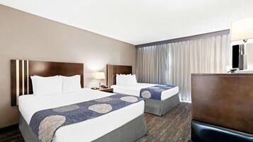 A hotel room with two beds, a desk, a lamp, and a large window with curtains.