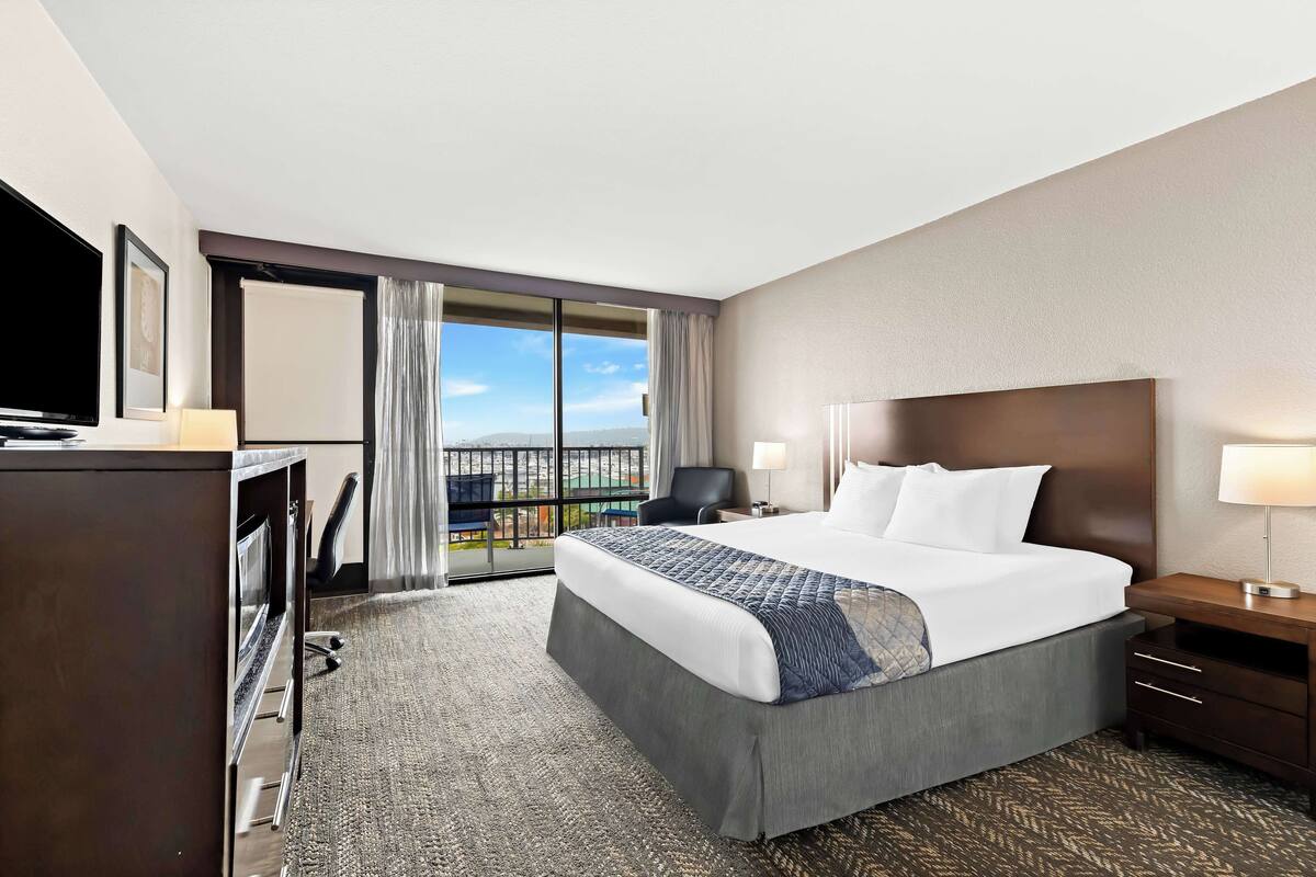 A hotel room with a large bed, a desk with a chair, a TV, and a view of the city through a large window.