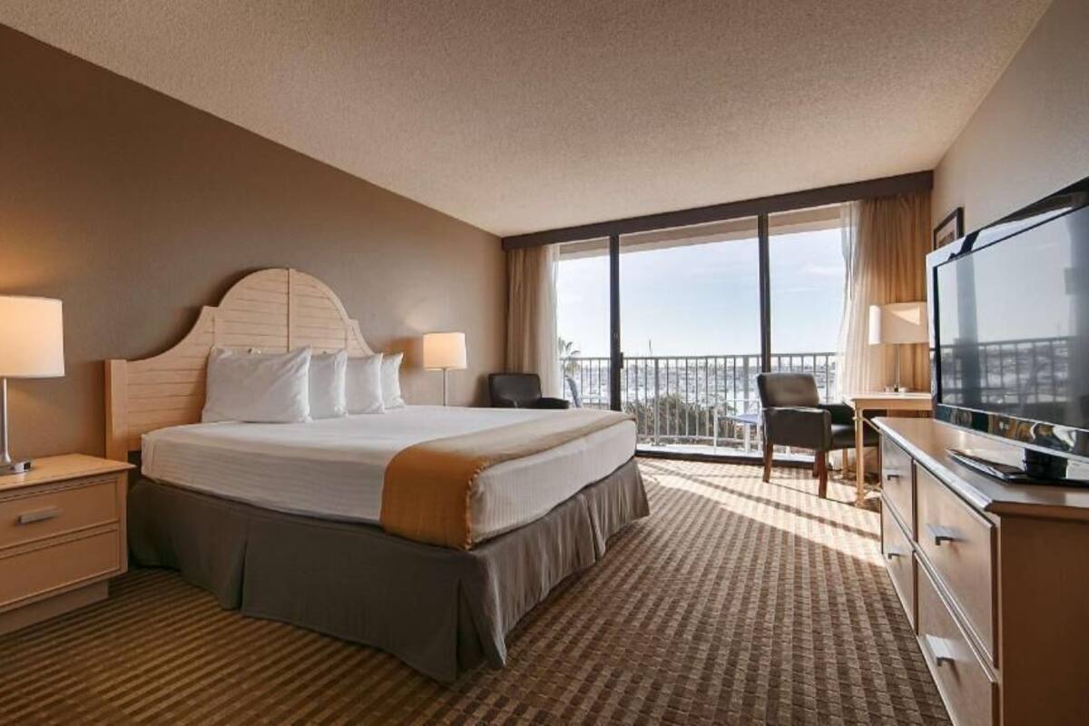 A hotel room with a large bed, a desk with a TV, a chair, and a balcony with a view.