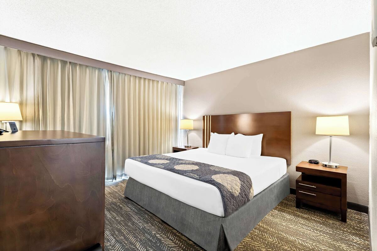 A hotel room with a bed, bedside tables, a desk, and a large window with curtains.