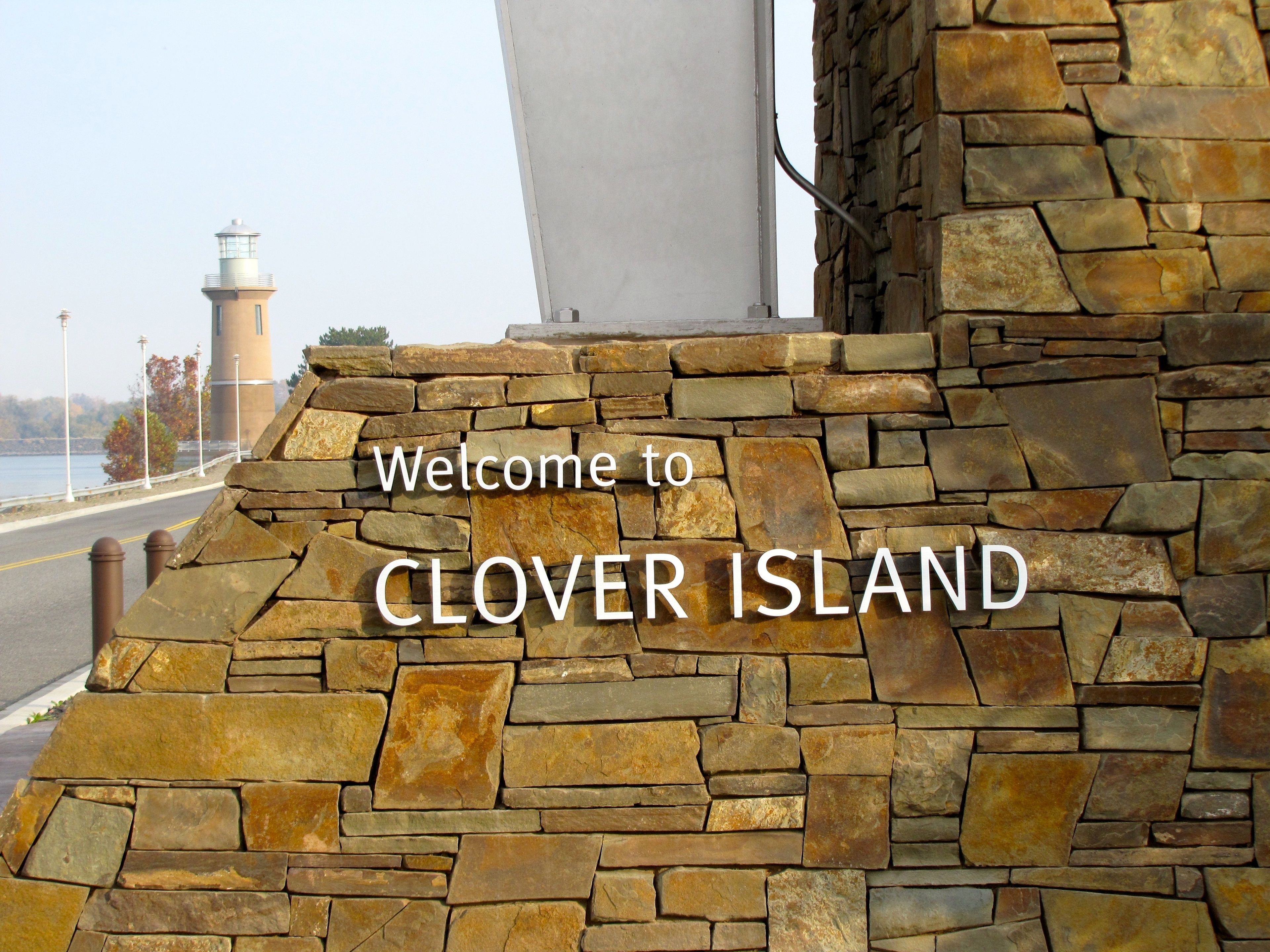 A stone wall with the text "Welcome to CLOVE ISLAND" and a lighthouse in the background.