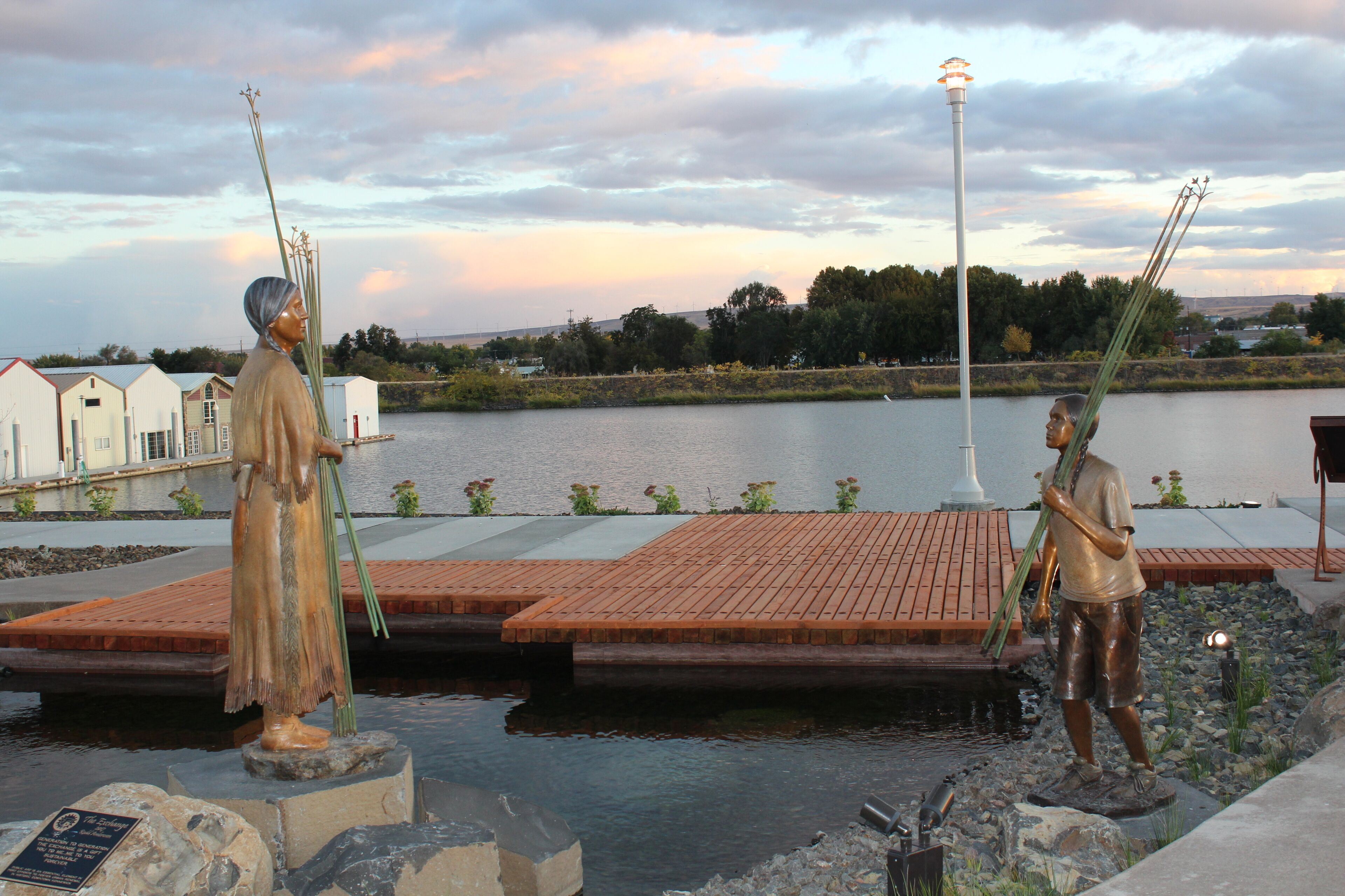 A statue of a person holding a spear by a body of water with a brick platform in the foreground.