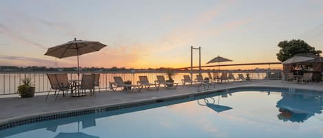 A swimming pool with lounge chairs and umbrellas, a view of the sunset, and a railing along the pool's edge.