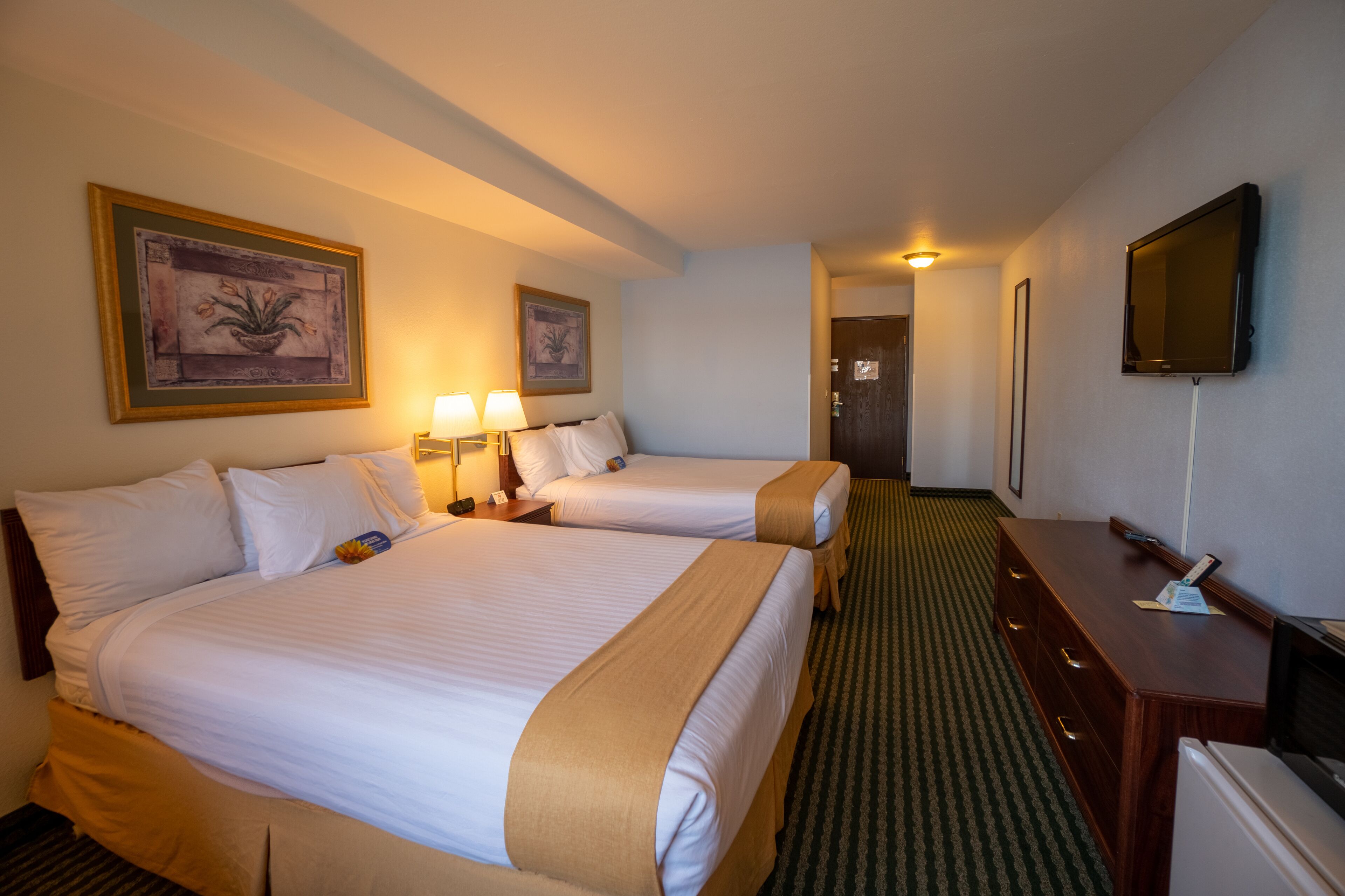 A hotel room with two beds, a desk, a television, and framed artwork on the walls.