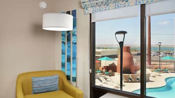 Un fauteuil jaune avec un coussin à rayures, une grande fenêtre donnant sur une piscine et un espace salon extérieur, et un climatiseur fixé au mur.