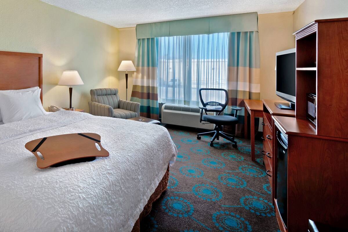 Une chambre d’hôtel avec un lit, un bureau avec un ordinateur, une chaise, une télévision et une fenêtre avec des rideaux.
