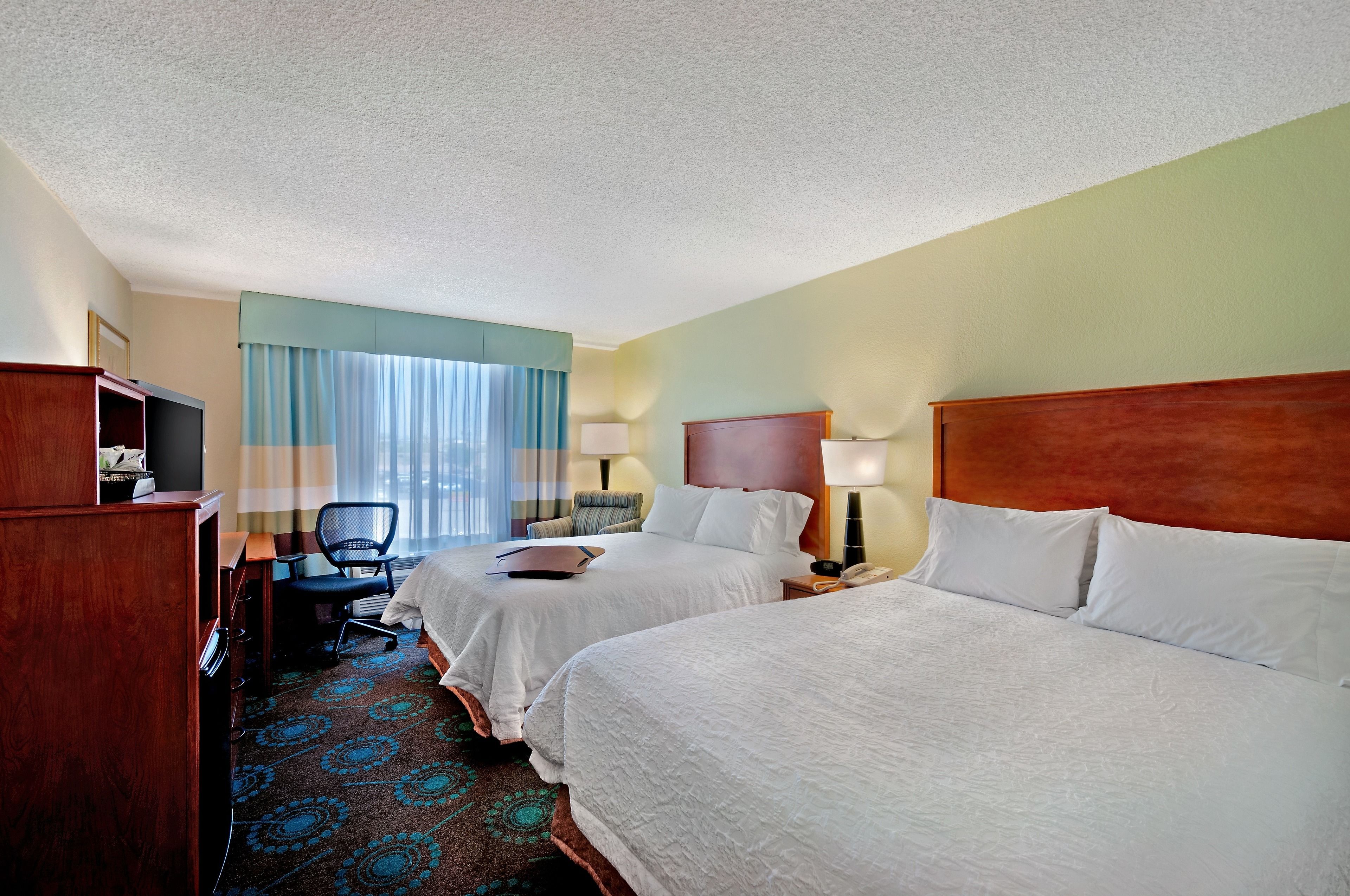 A hotel room with two beds, a desk, a chair, a TV, and a window with curtains.