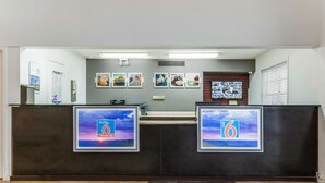 A reception desk with two monitors displaying the number 6, a bulletin board with various notices, and a window with blinds. - Motel 6 Alexandria, LA - South (Alexandria)