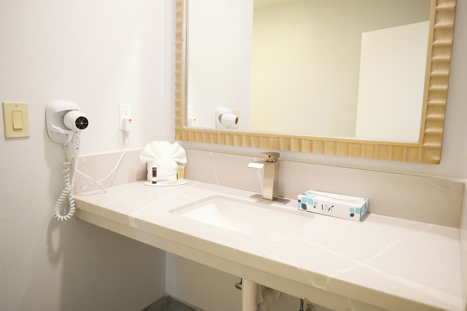 Et badeværelse med en hvid vask, en hårtørrer, et spejl og en æske med toiletartikler.