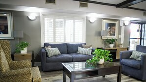 A living room with a dark grey sofa, patterned armchair, and a coffee table. There are two potted plants, a lamp, and a small table with decorative items. - Days Inn by Wyndham West Covina (West Covina)