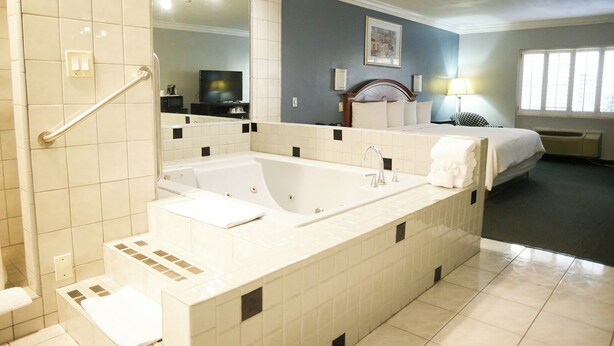 A modern bathroom with a large bathtub, a walk-in shower with a handrail, a vanity with a mirror, and a bed in the background. - Days Inn by Wyndham West Covina (West Covina)