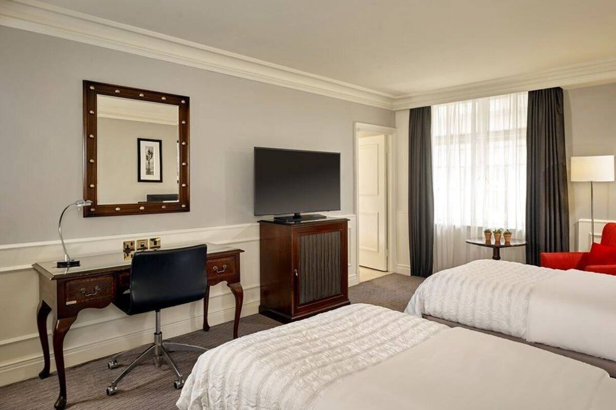 A hotel room with two beds, a TV, a desk with a chair, a mirror, and a red chair.