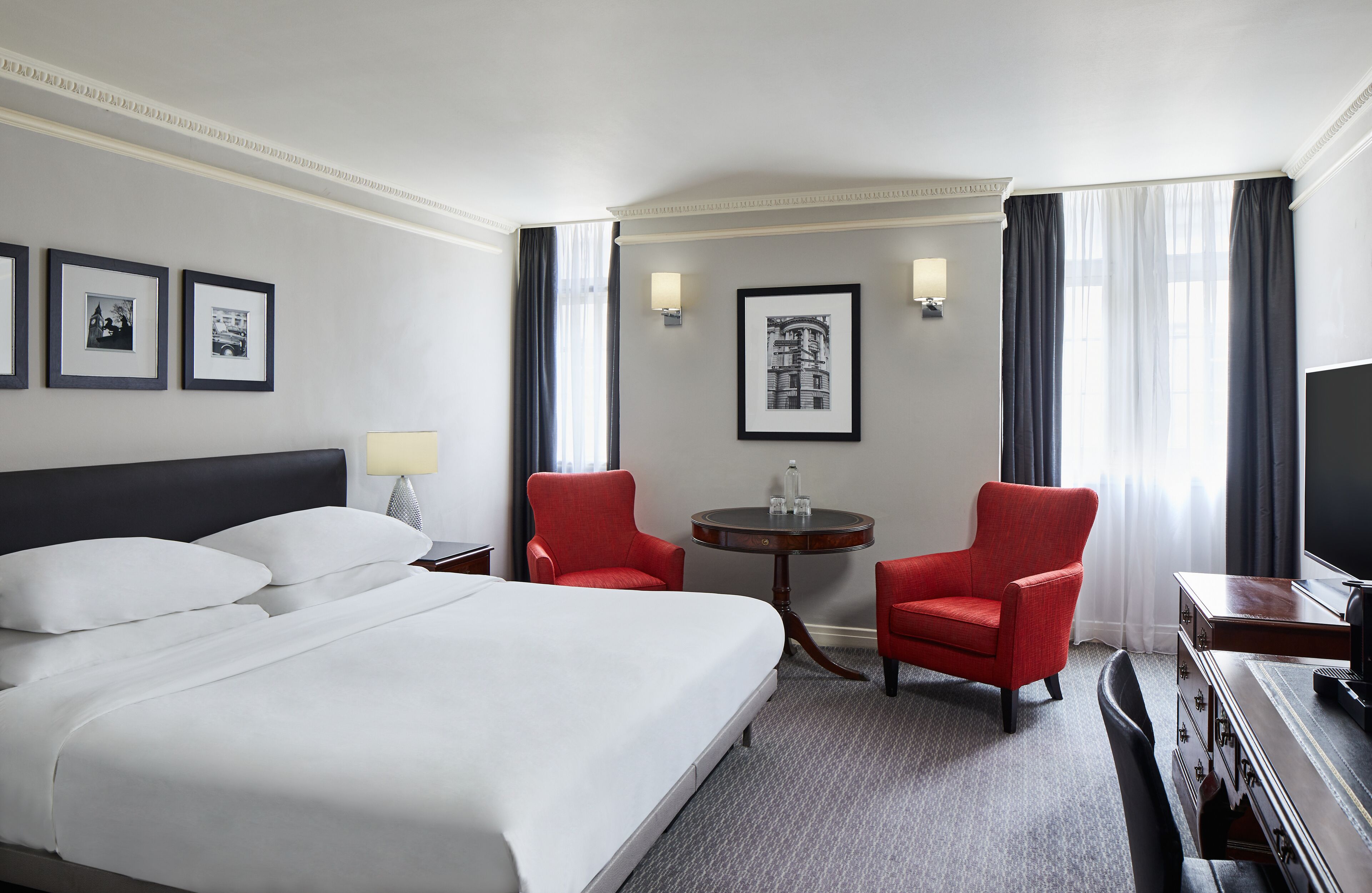 A hotel room with a large bed, two red armchairs, a small table, a TV, and framed pictures on the wall.