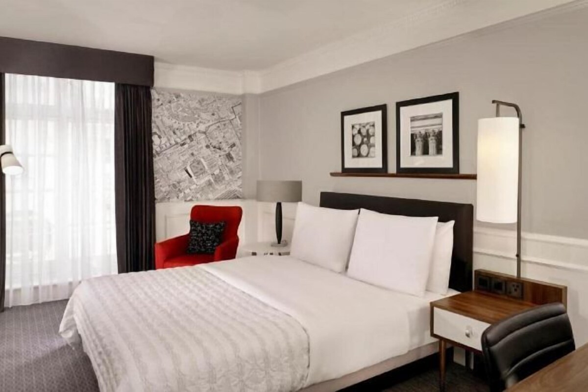 A hotel room with a large bed, a nightstand, a red chair, and a wall-mounted lamp.