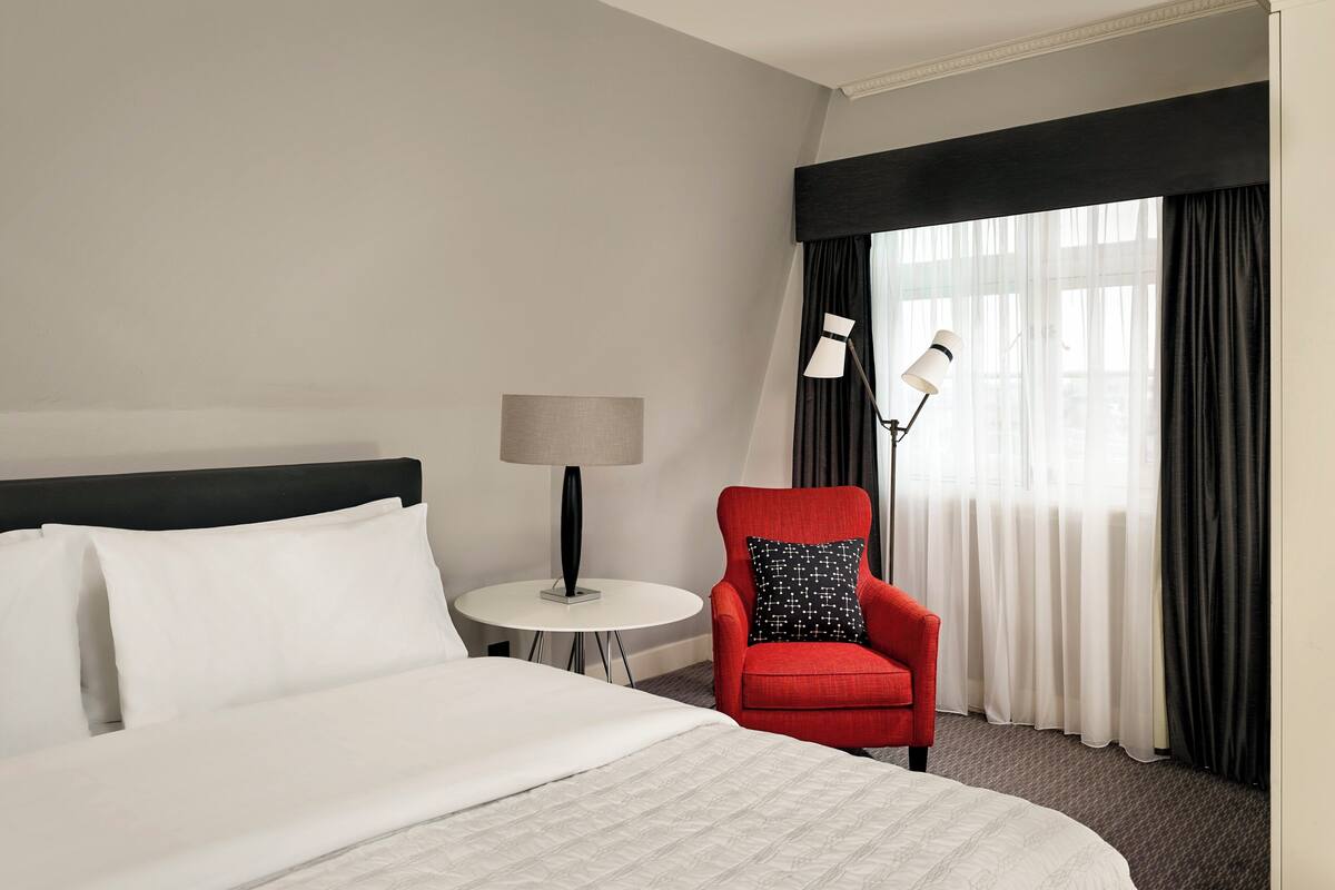 A hotel room with a bed, a red armchair, a small round table with a lamp, and a large window with curtains.