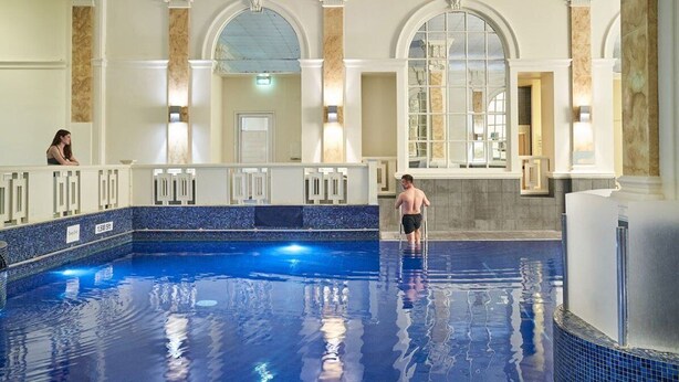 A large indoor swimming pool with a tiled floor and walls, surrounded by a poolside area with a railing.