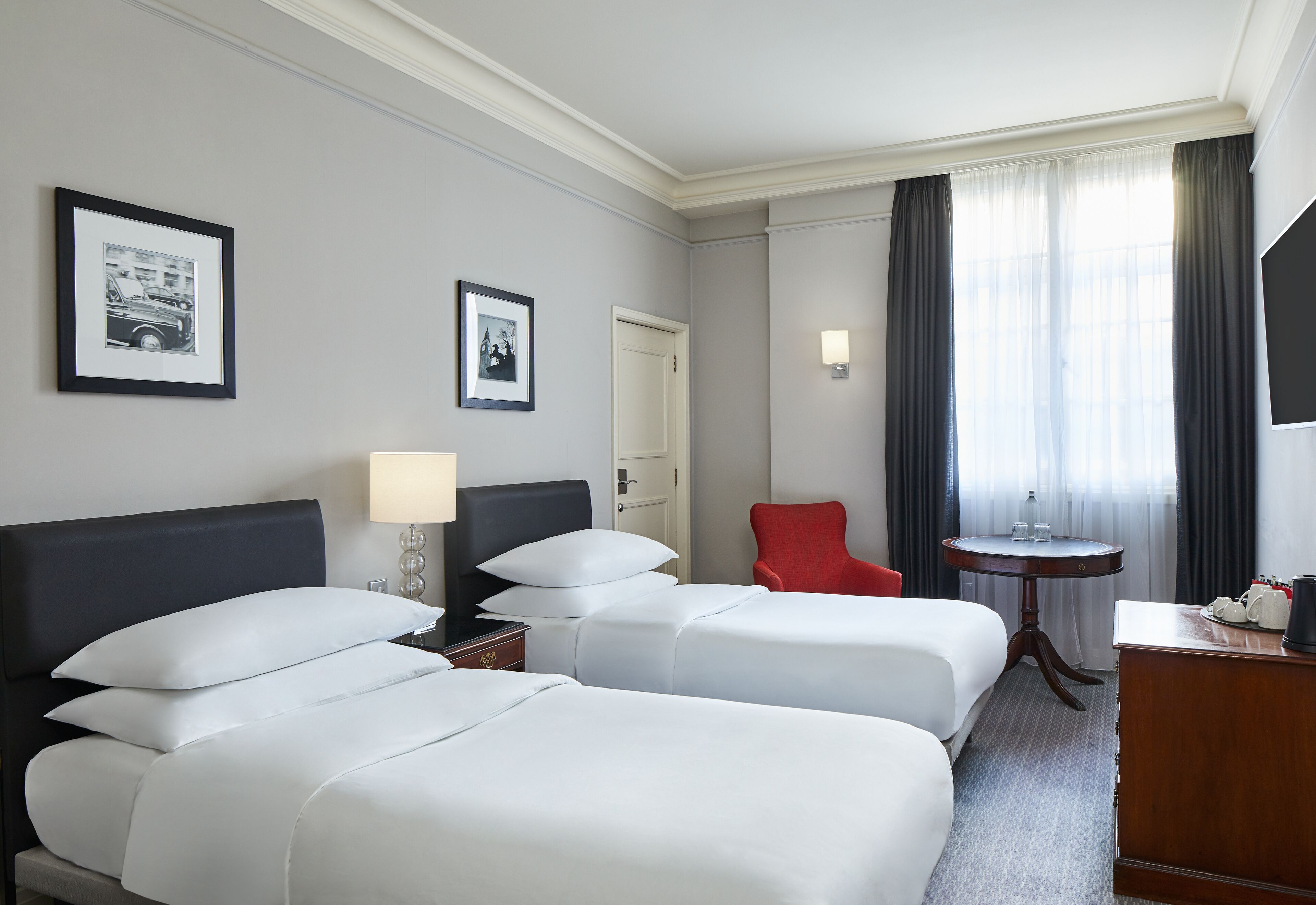 A hotel room with two beds, a red chair, a small table with a lamp, and framed pictures on the wall.