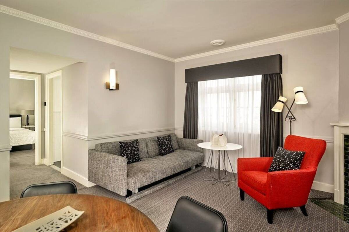A hotel room with a grey sofa, a red armchair, a small round table, and a fireplace.
