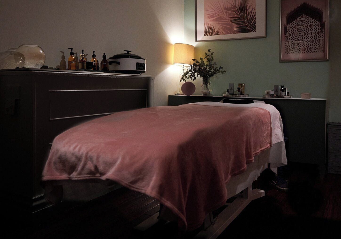 A dimly lit room with a bed, a dresser, and a nightstand. There is a lamp on the nightstand, a vase with a plant, and a framed picture on the wall.