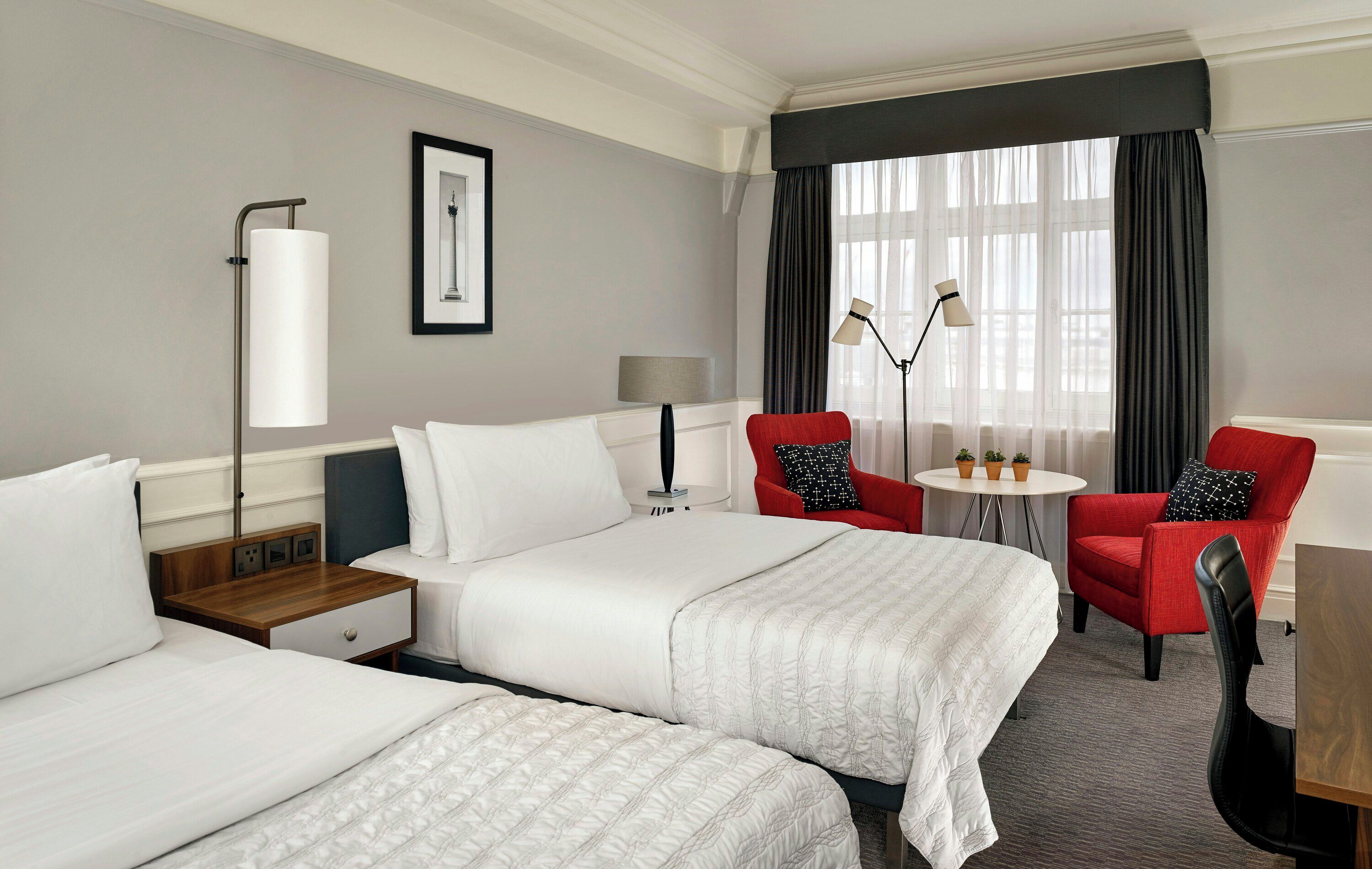 A hotel room with two beds, a desk, a chair, a lamp, and a window with curtains.