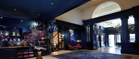 A spacious lobby with a large dark blue carpet, a reception desk with a vase of flowers, and a view of the city through a large arched doorway.