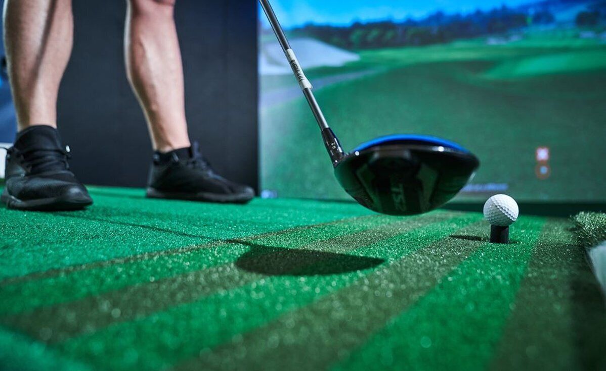 A golf club positioned over a golf ball on a green mat, with a golfer's feet visible in the background.