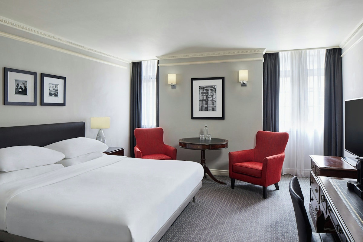 A hotel room with a large bed, two red armchairs, a small table, a television, and framed pictures on the wall.